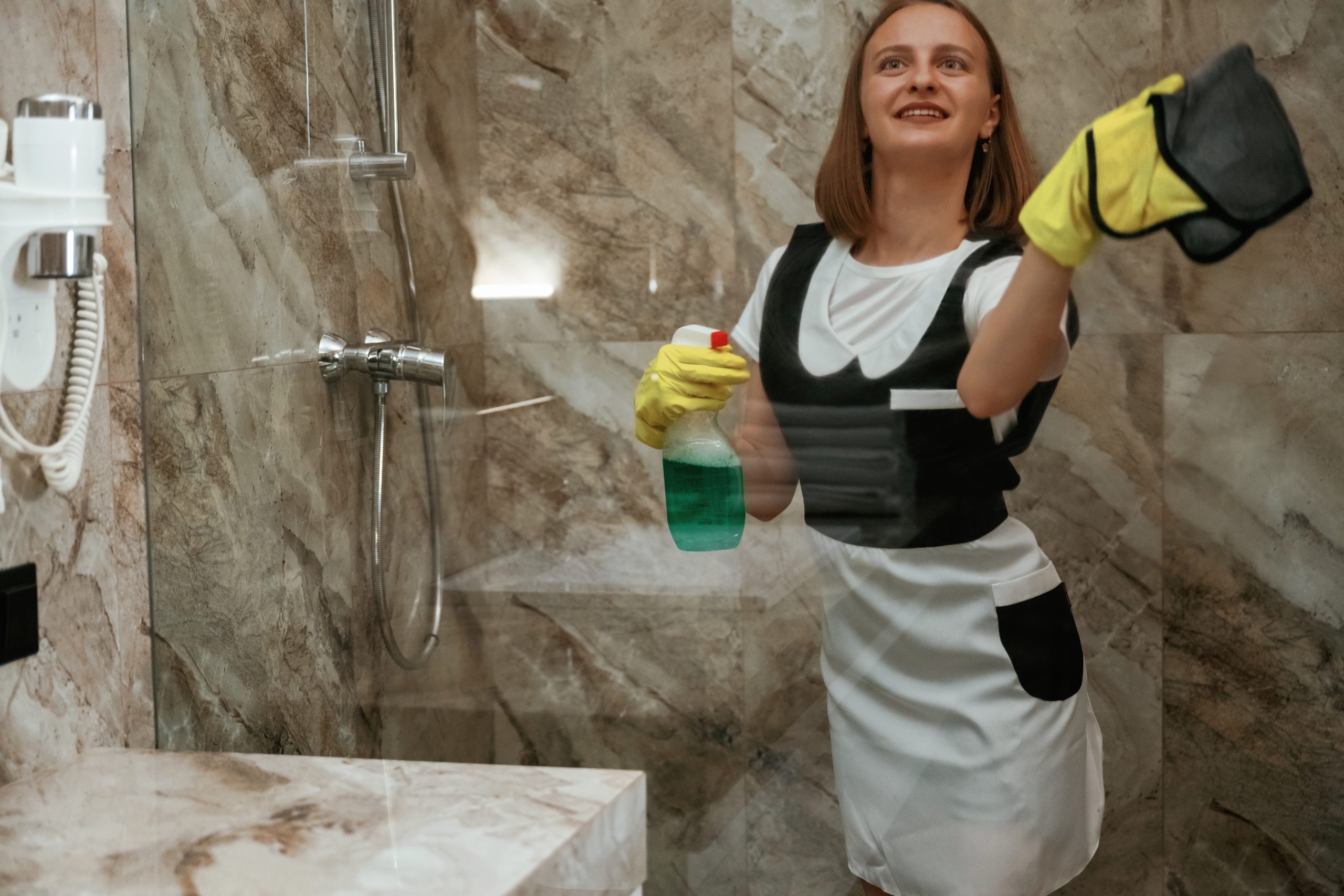 Green washing product, cleaning the glass in bathroom. Young chambermaid is in hotel room.