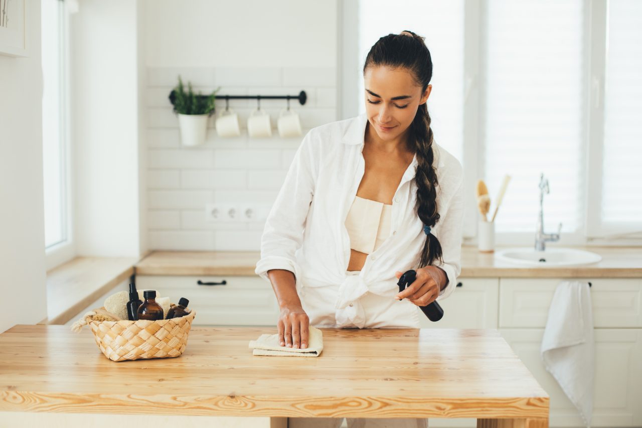 Young woman cleaning wooden table using spray in glass bottle and natural rag in a kitchen.