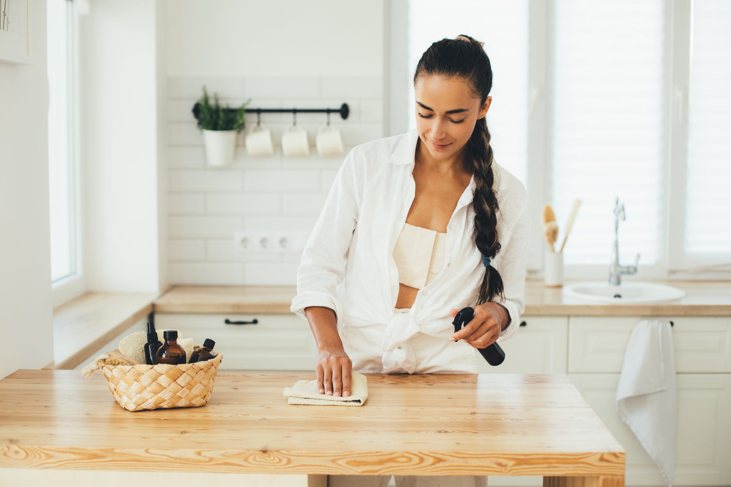 Young woman cleaning wooden table using spray in glass bottle and natural rag in a kitchen.